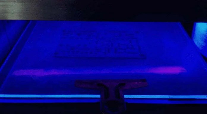 A high-contrast shot of a flat substrate under intense blue-violet ultraviolet light, showing a faint rectangular circuit pattern and a squeegee tool at the bottom edge.