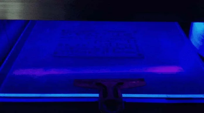 IC Reverse Engineering 13 A high-contrast shot of a flat substrate under intense blue-violet ultraviolet light, showing a faint rectangular circuit pattern and a squeegee tool at the bottom edge.